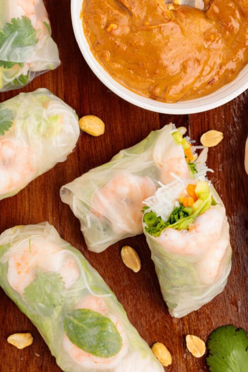 Vietnamese Shrimp Salad Rolls on a white plate with fresh herbs around it and a small bowl of peanut dipping sauce.
