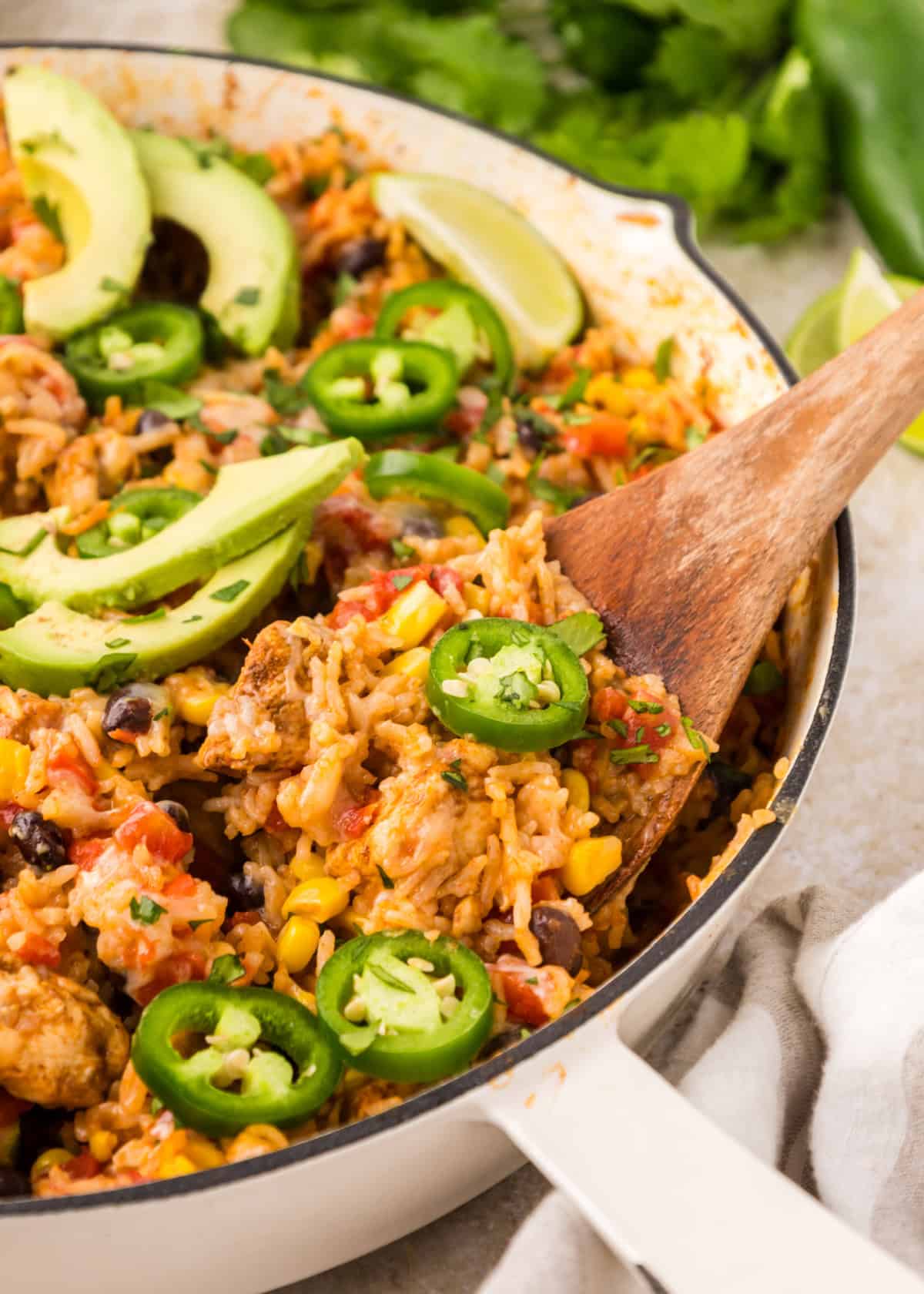 Southwest Chicken Skillet in a pan with a wooden spoon with fresh avocado slices and sliced jalapeños on top.