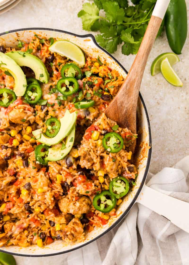 Southwest Chicken Skillet in a pan with a wooden spoon with fresh avocado slices and sliced jalapeños on top.