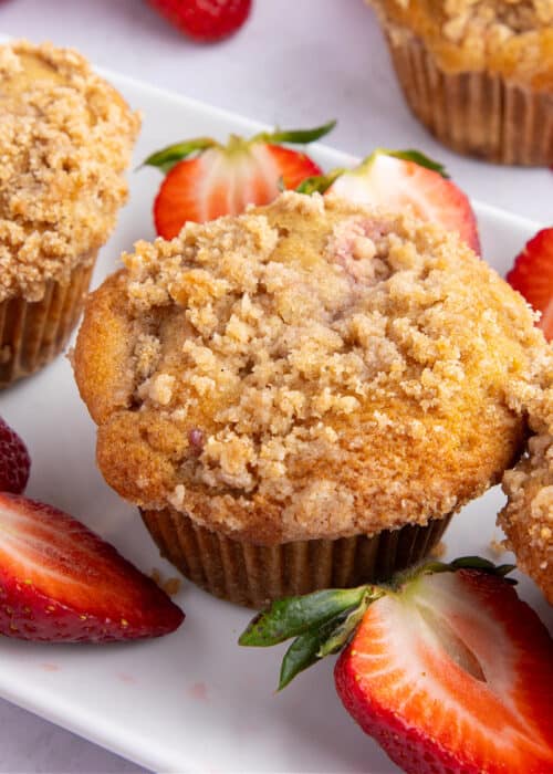 Strawberry rhubarb muffins on a white plate with fresh strawberry slices beside them.