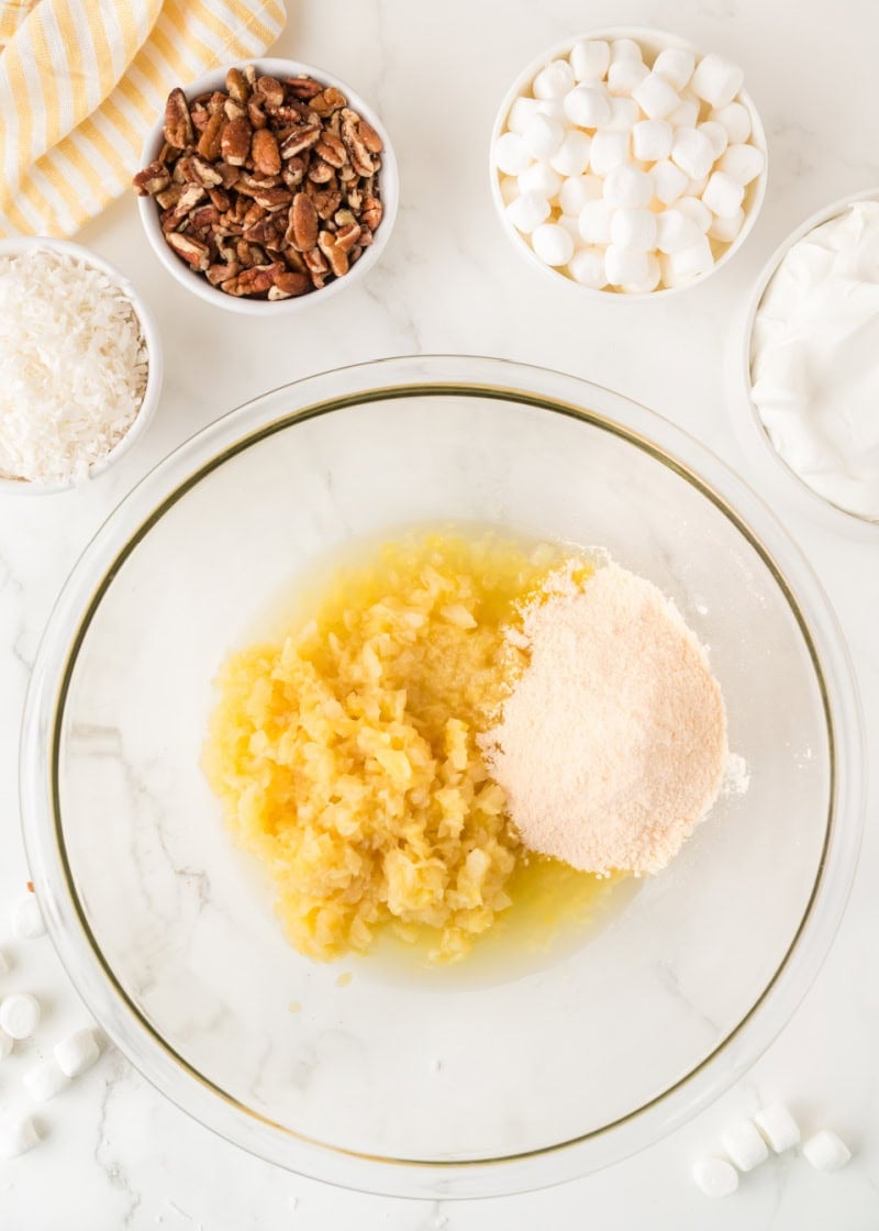 crushed pineapple and vanilla pudding mix in a clear bowl beside bowls of marshmallows, coconut, pecans and whipped topping.