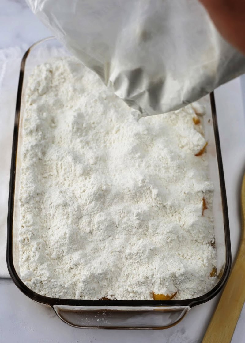 pouring cake mix over a peaches in a glass casserole dish.
