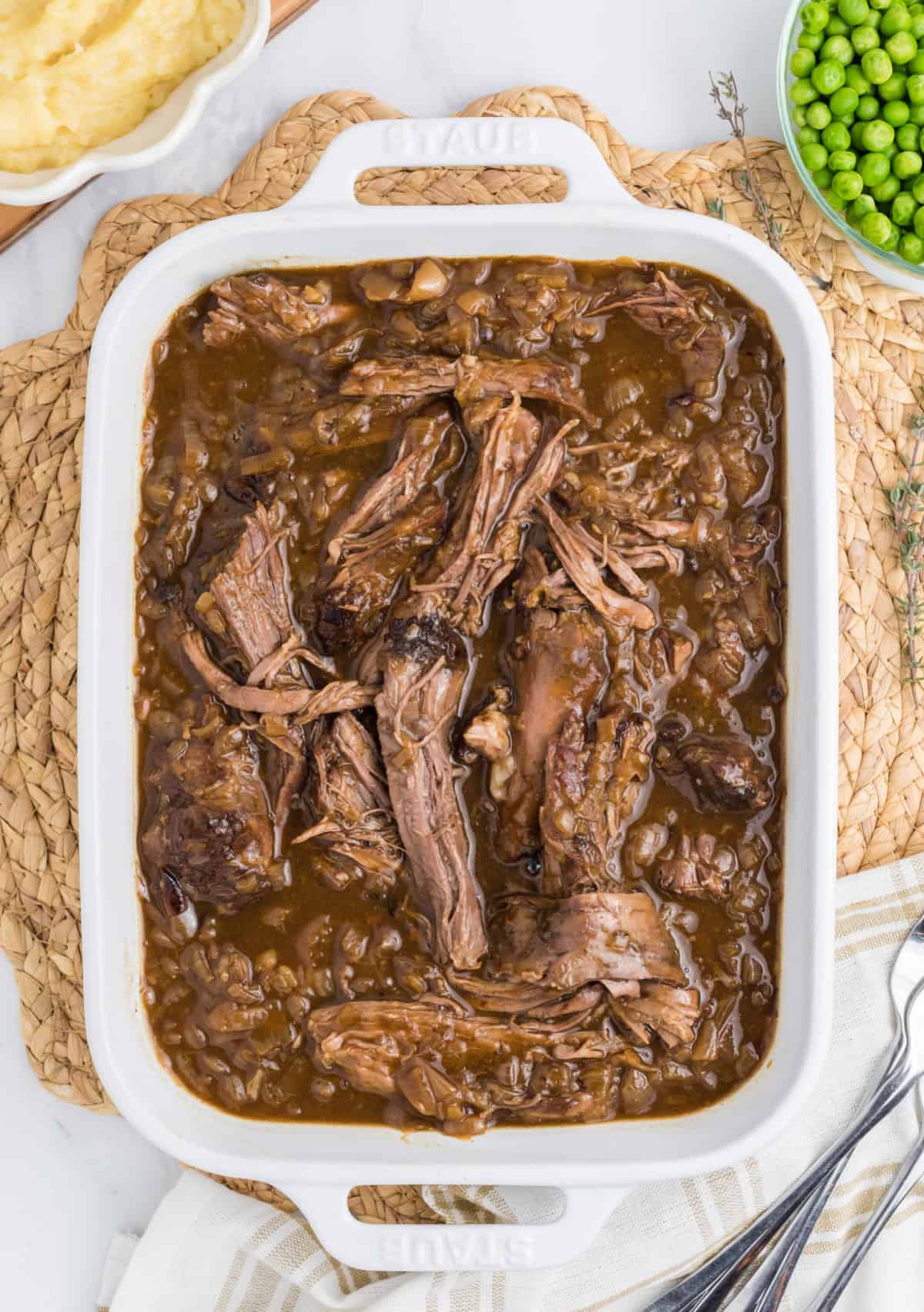 a casserole dish with shredded beef roast with onions and a bowl of mashed potatoes and peas beside it.