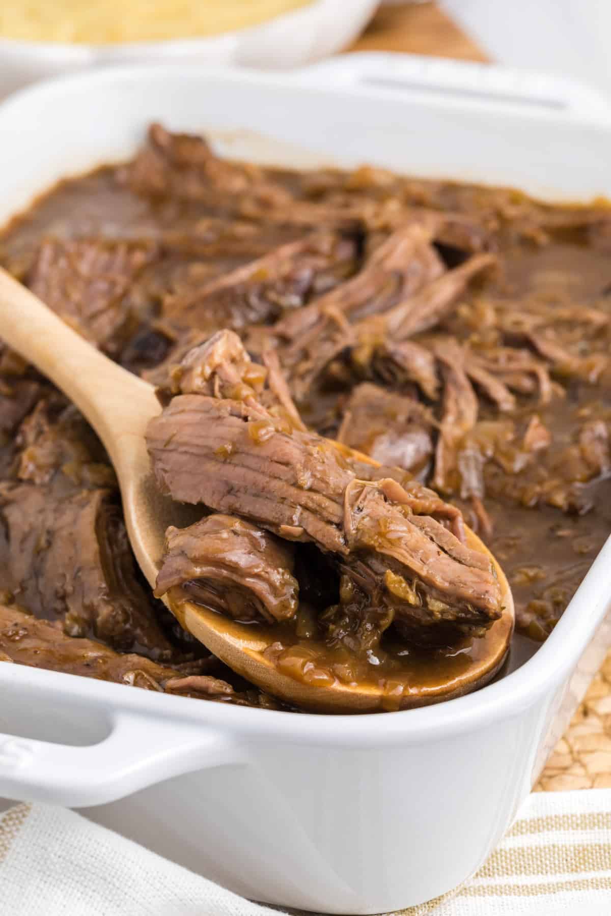 a casserole dish with shredded beef roast and a wooden spoon.
