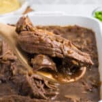 a casserole dish with shredded beef roast with onions and a wooden spoon.
