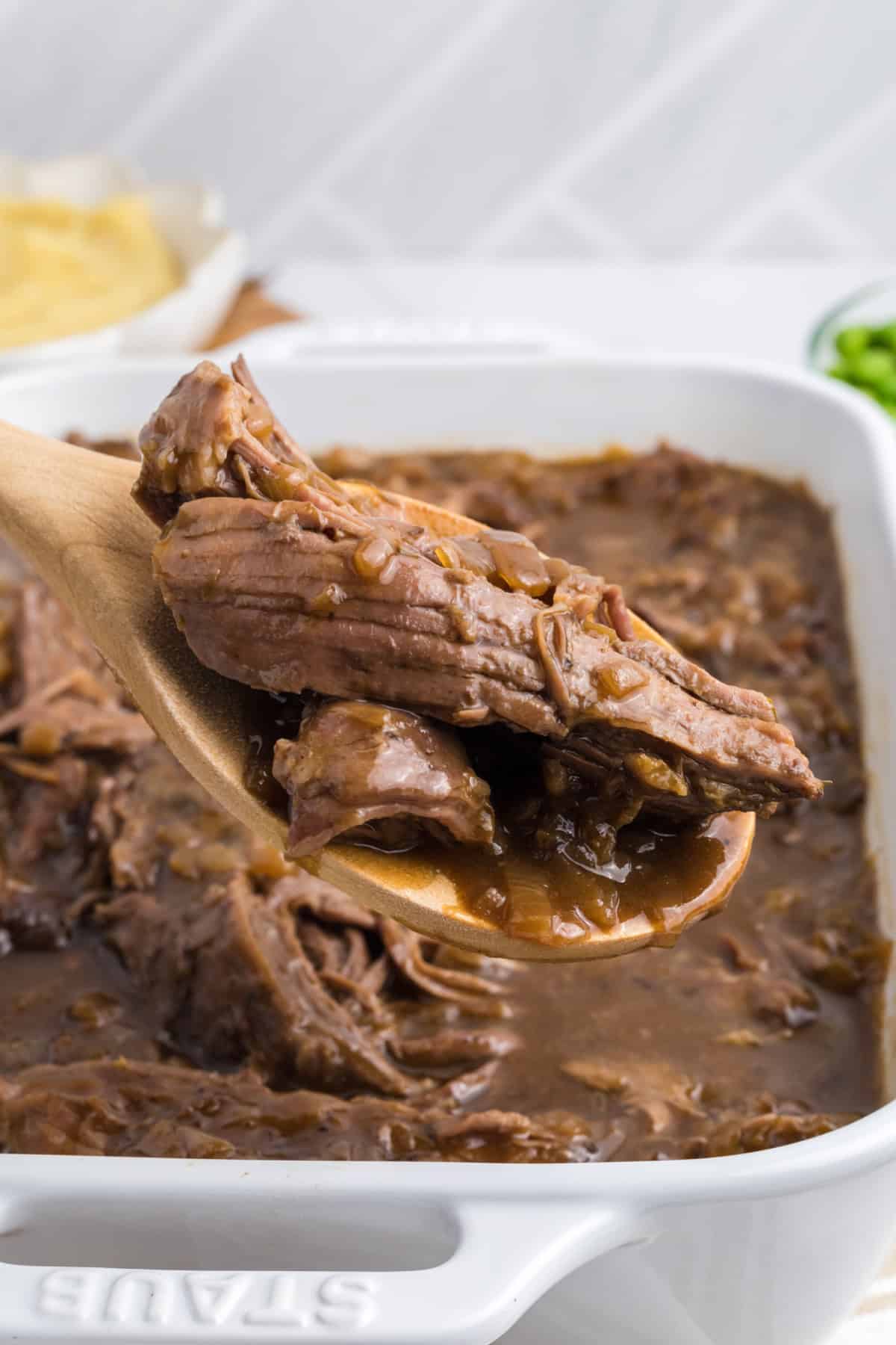 a casserole dish with shredded beef roast with onions and a wooden spoon.