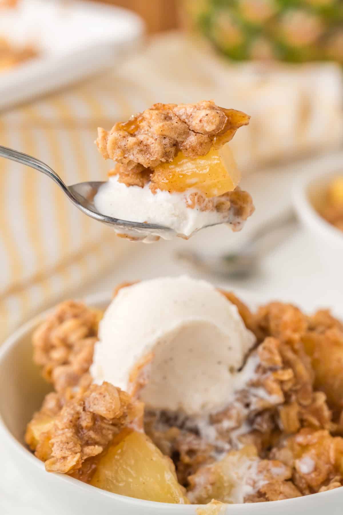 Pineapple crisp in a bowl with a scoop of vanilla ice cream, and a spoon lifting up some.