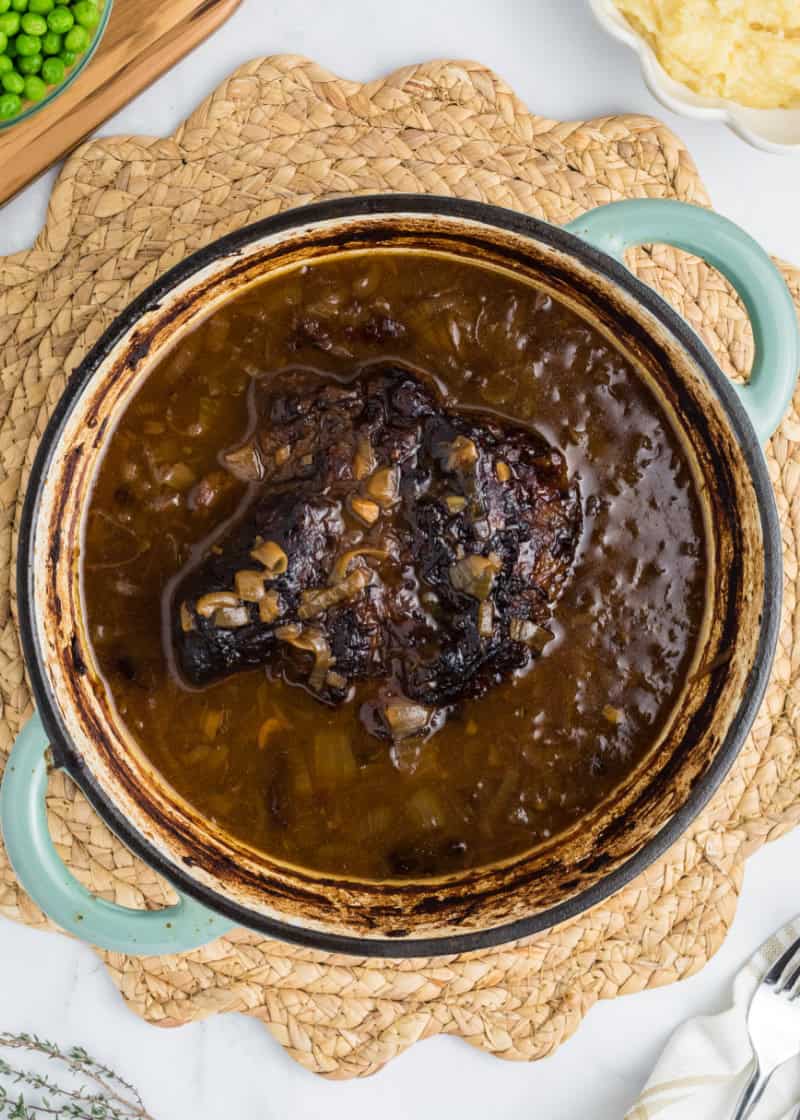 A dutch oven with a french onion pot roast inside with peas and potatoes in bowls beside it.
