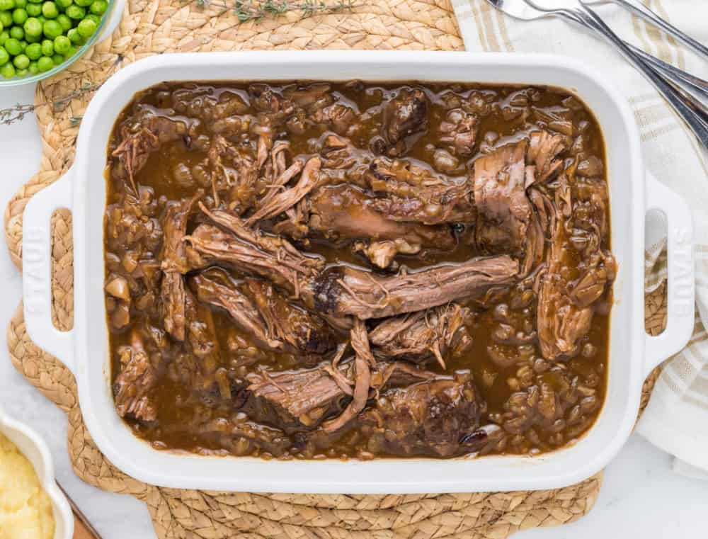 A white casserole dish filled with french onion beef roast and peas on the side in a bowl.
