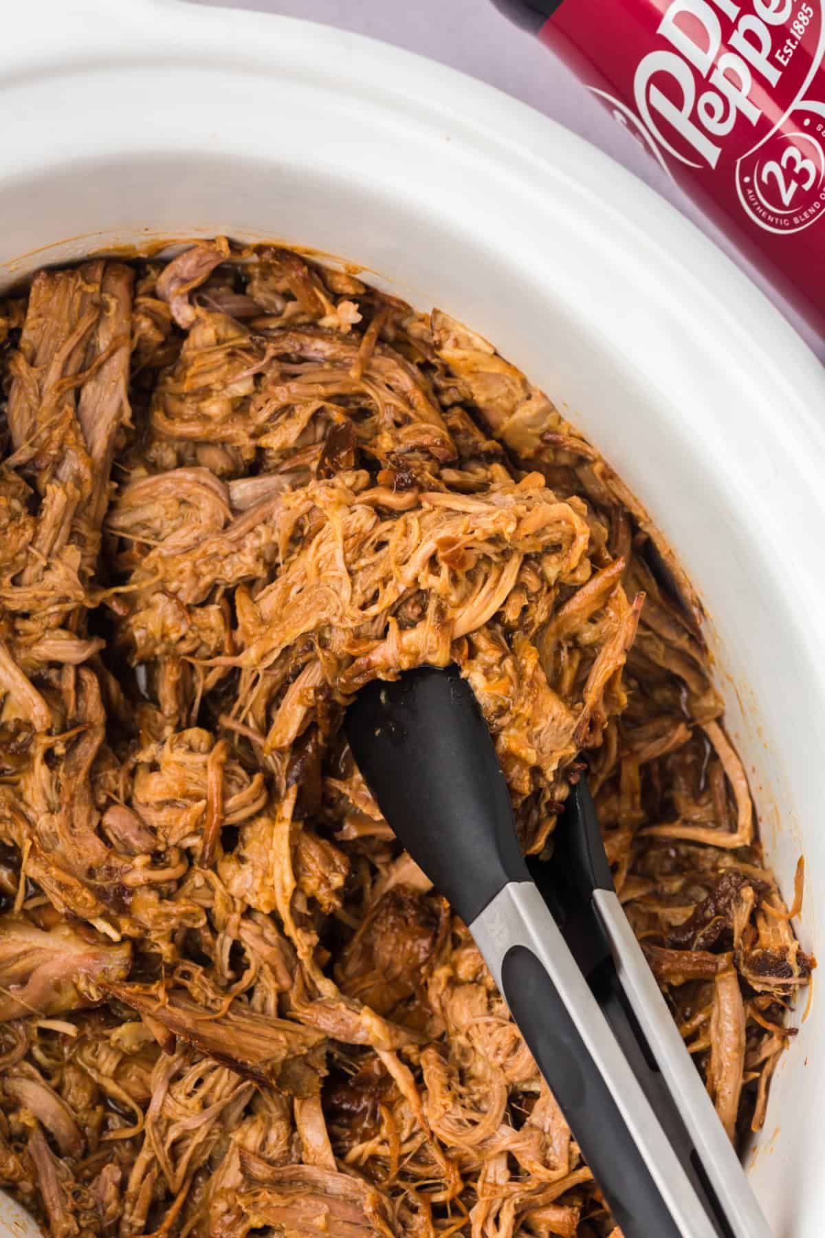 Slow cooker pulled pork being lifted out of a crockpot using tongs.