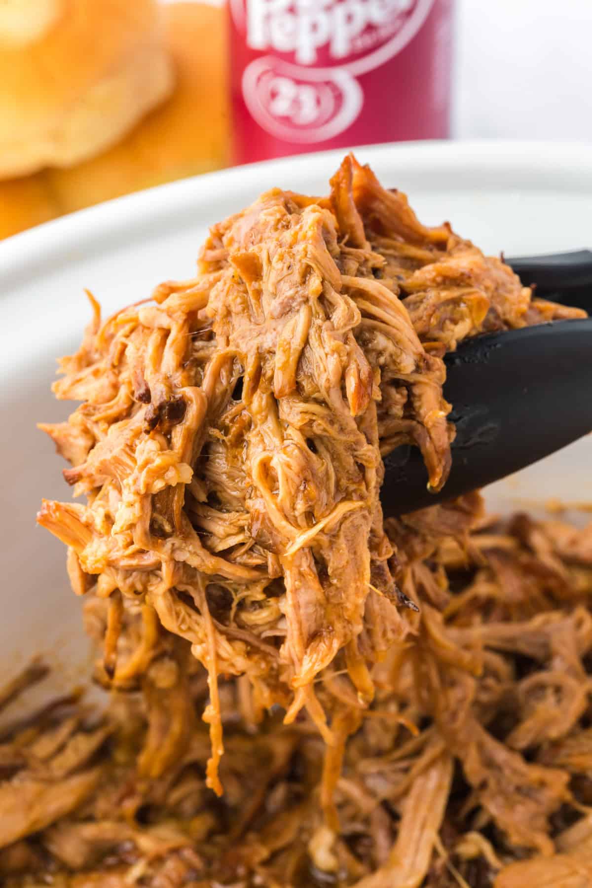 Slow cooker pulled pork being lifted out of a crockpot using tongs with a bottle of dr. pepper in the background.