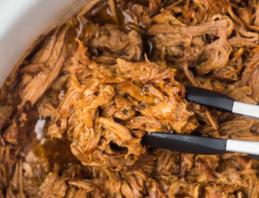 pulled pork in a crockpot with black tongs.