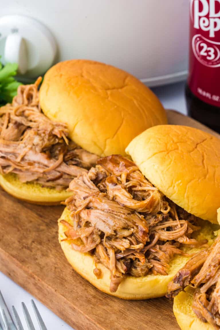 pulled pork on sandwich buns in front of a slow cooker and a bottle of soda.