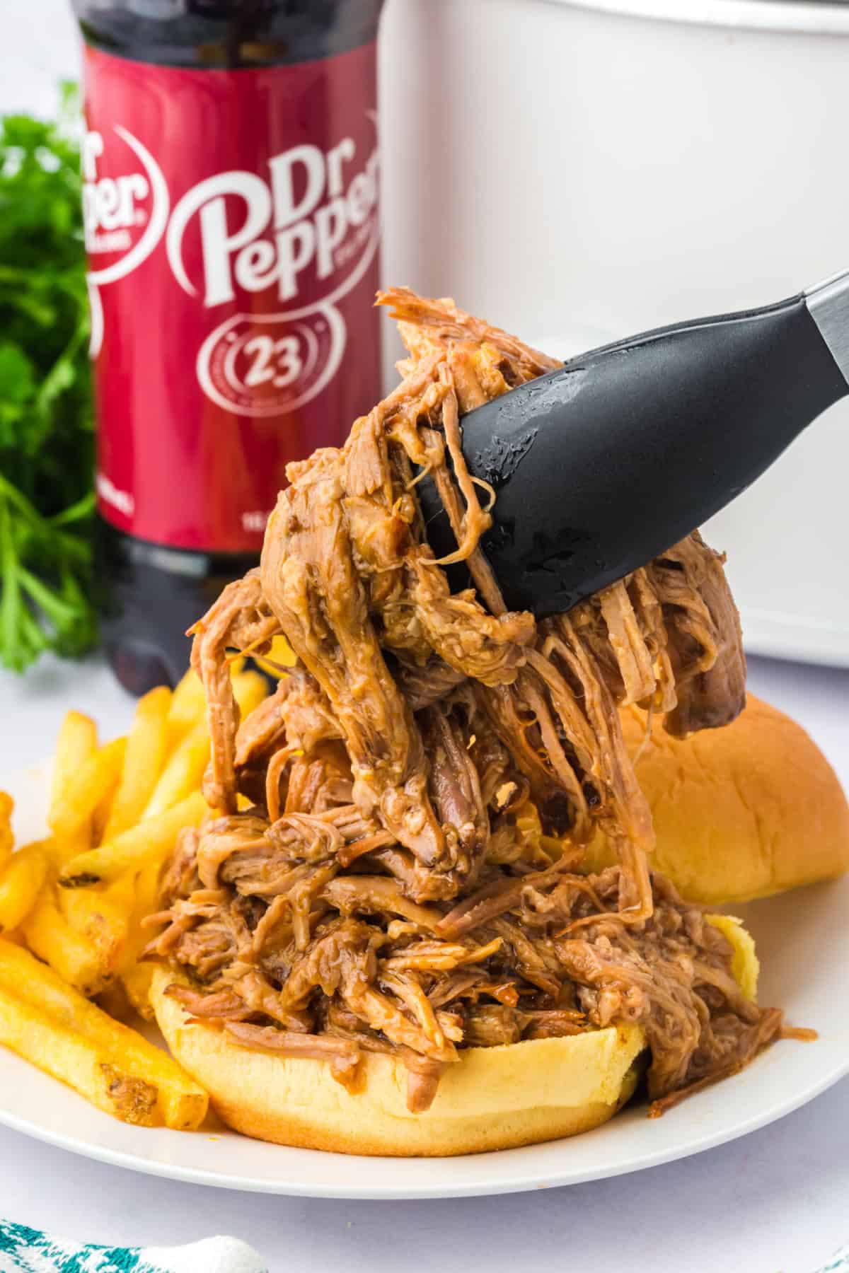 Slow cooker pulled pork being put in a sandwich bun using tongs with a crockpot and a bottle of dr. pepper soda in the background.