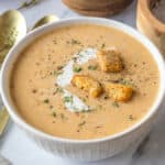 A white bowl of Butternut Squash Soup with Apple with a swirl of cream, croutons, fresh herbs, and black pepper.