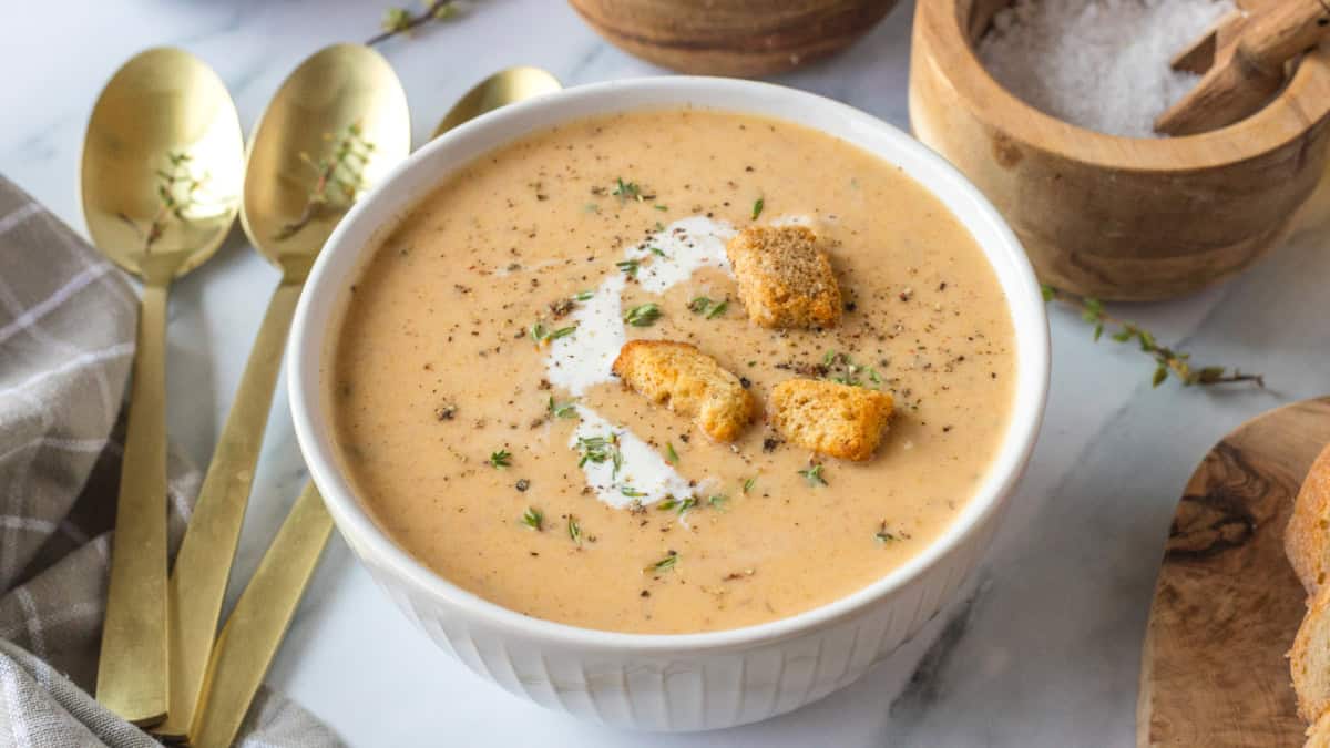 A white bowl of Butternut Squash Soup with Apple with a swirl of cream, croutons, fresh herbs, and black pepper.