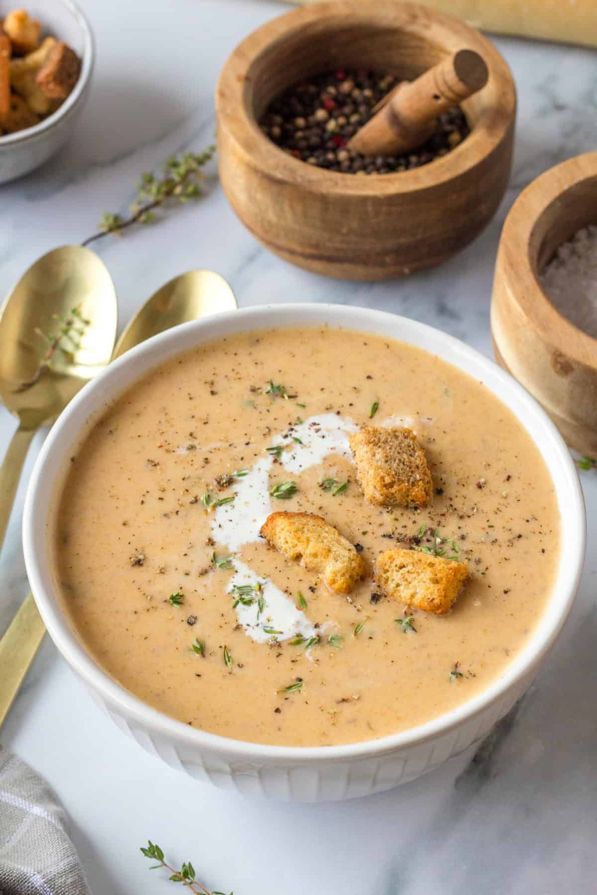 A white bowl of Butternut Squash Apple Soup with a swirl of cream, croutons, fresh herbs, and black pepper.