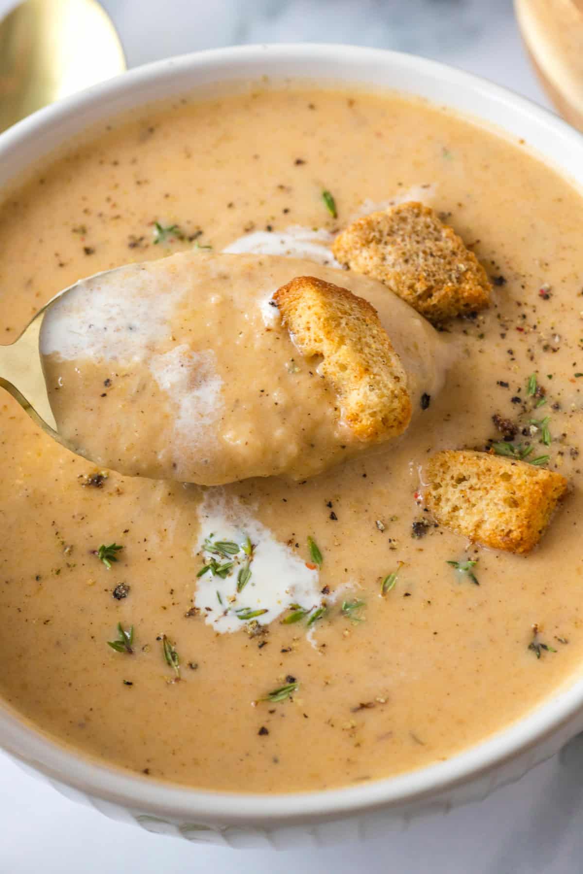 A white bowl of Apple Butternut Squash Soup with a swirl of cream, croutons, fresh herbs, and black pepper.