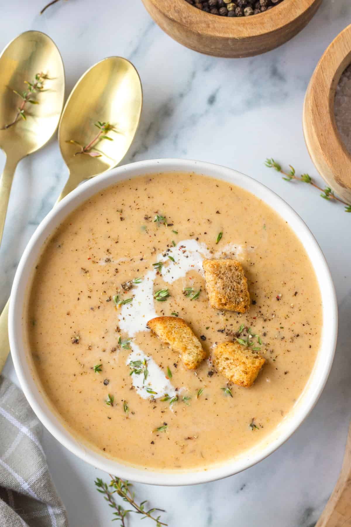 A white bowl of Butternut Squash Soup with Apple with a swirl of cream, croutons, fresh herbs, and black pepper.
