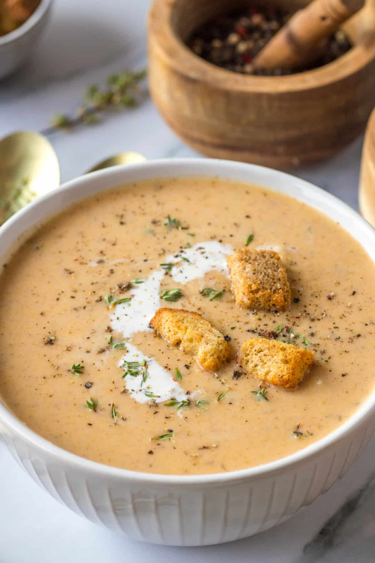 A white bowl of Butternut Squash Soup with Apple with a swirl of cream, croutons, fresh herbs, and black pepper.