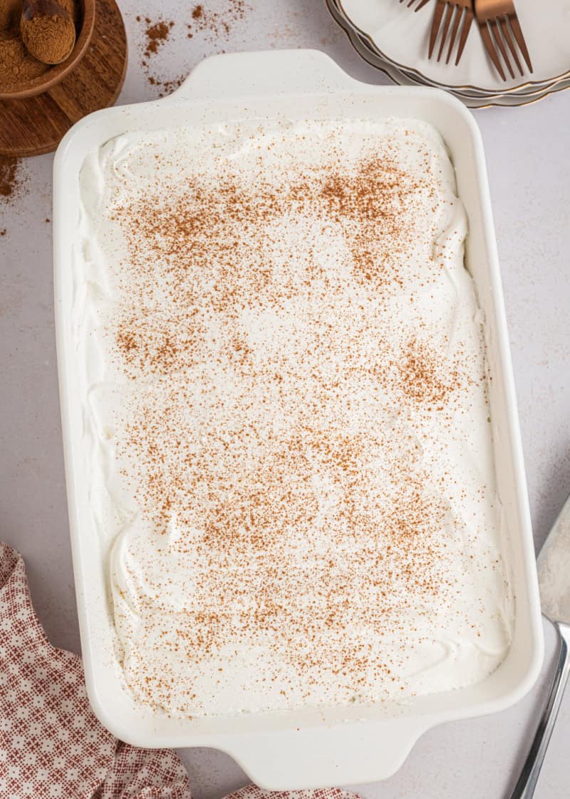A whole eggnog poke cake with whipped topping and cinnamon on top in a white rectangle baking dish.