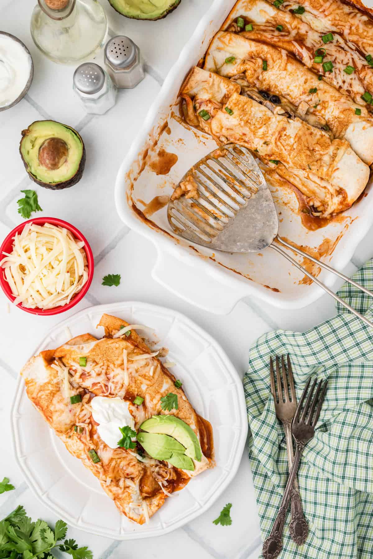 Turkey Enchiladas on a plate and in a baking dish with sides of avocado and grated cheese.