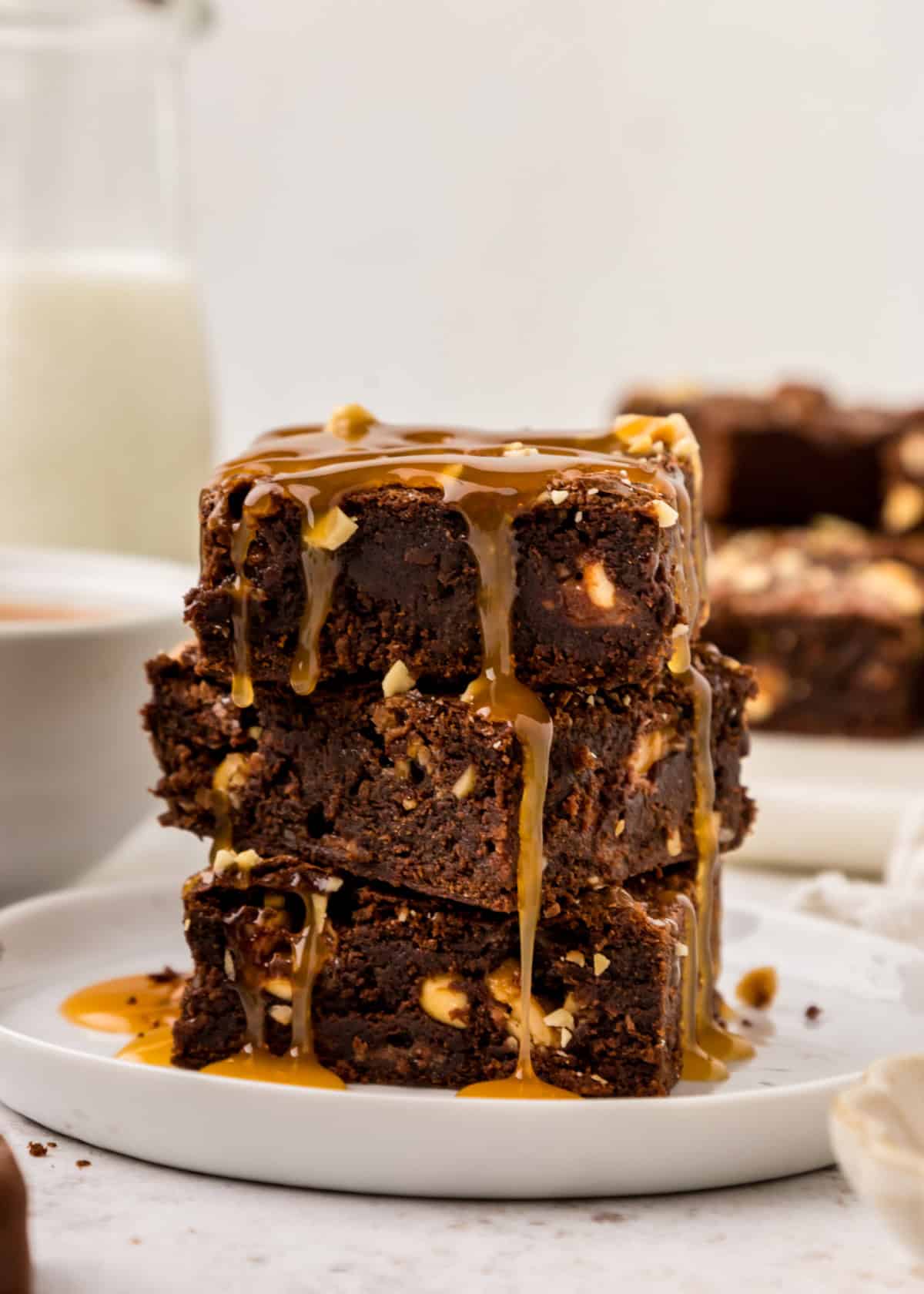 Snickers Brownies stacked on a white plate with caramel sauce drizzled on top.