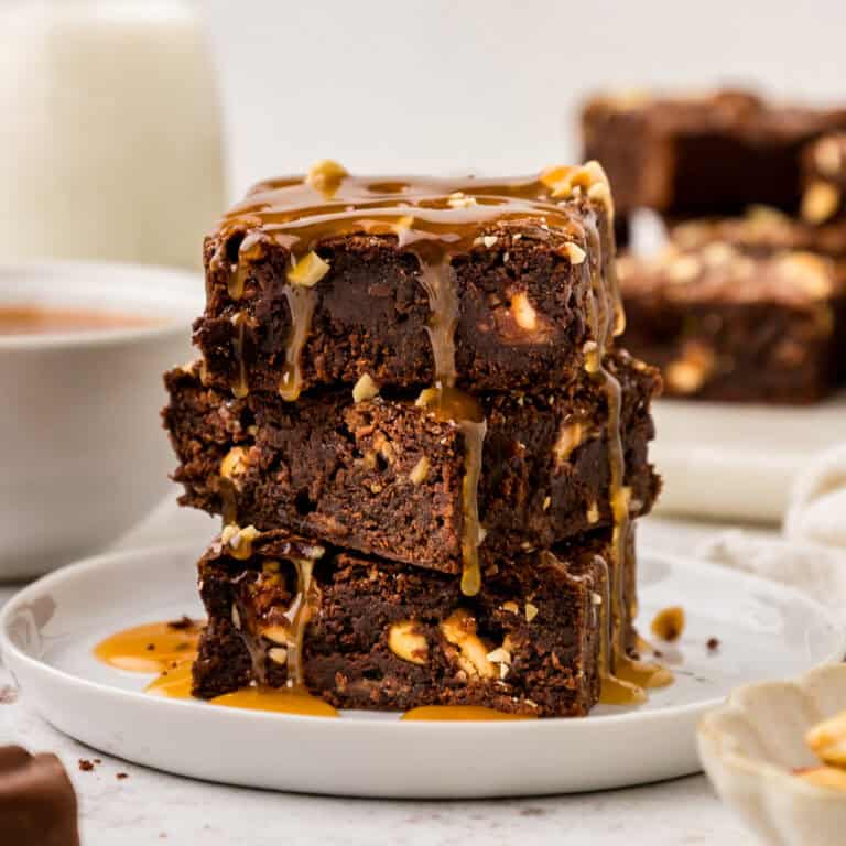 Snickers Brownies stacked on a white plate with caramel sauce drizzled on top.
