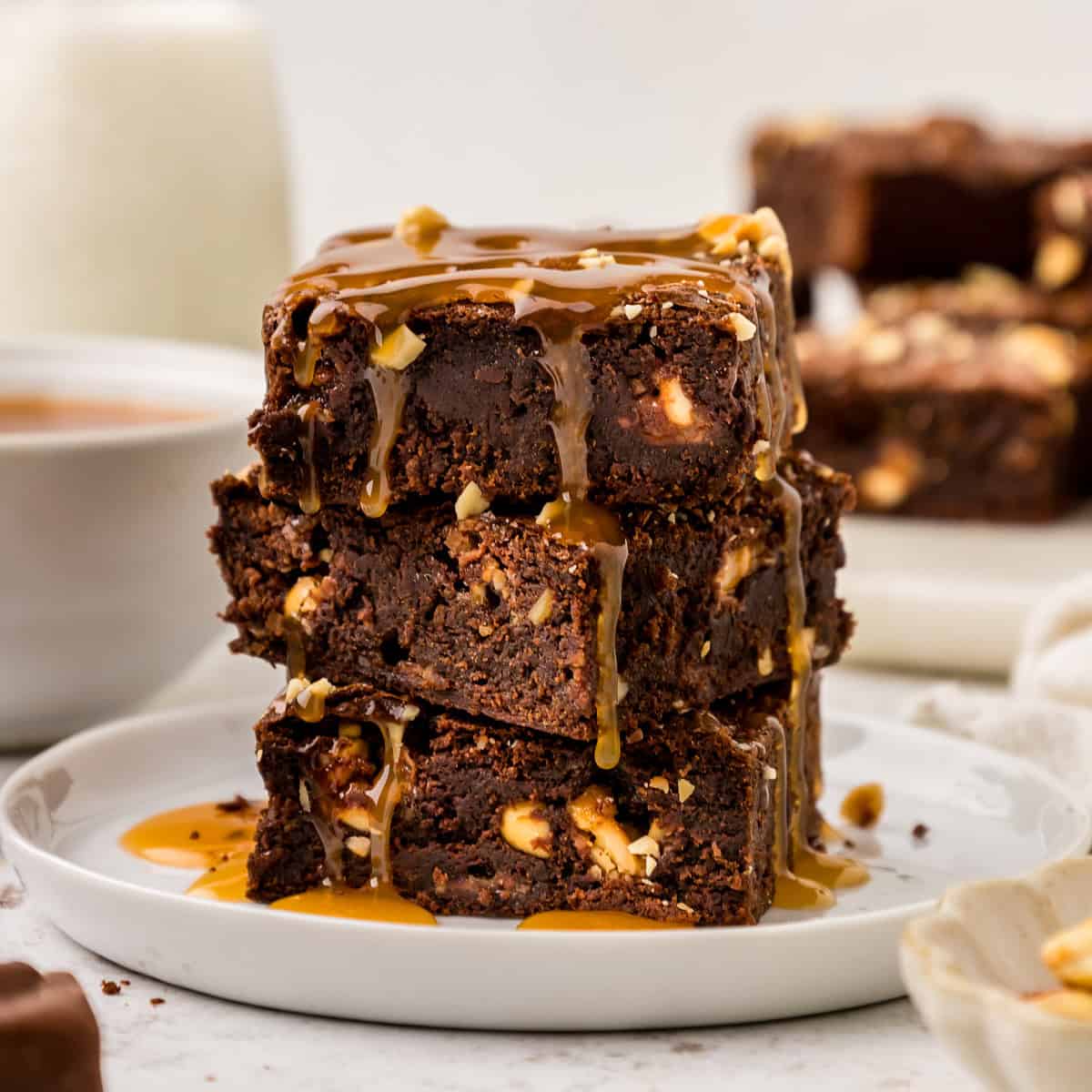 Snickers Brownies stacked on a white plate with caramel sauce drizzled on top.