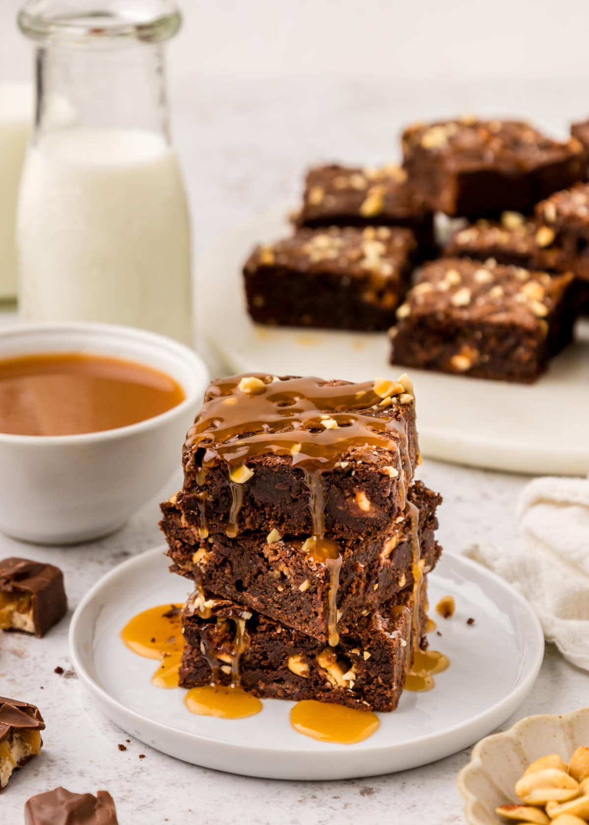 Snickers Brownies stacked on a white plate with caramel sauce drizzled on top.