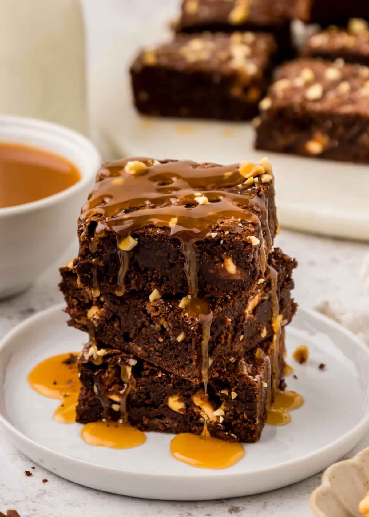 Snickers Brownies in a pan and stacked on a white plate with caramel sauce drizzled on top.