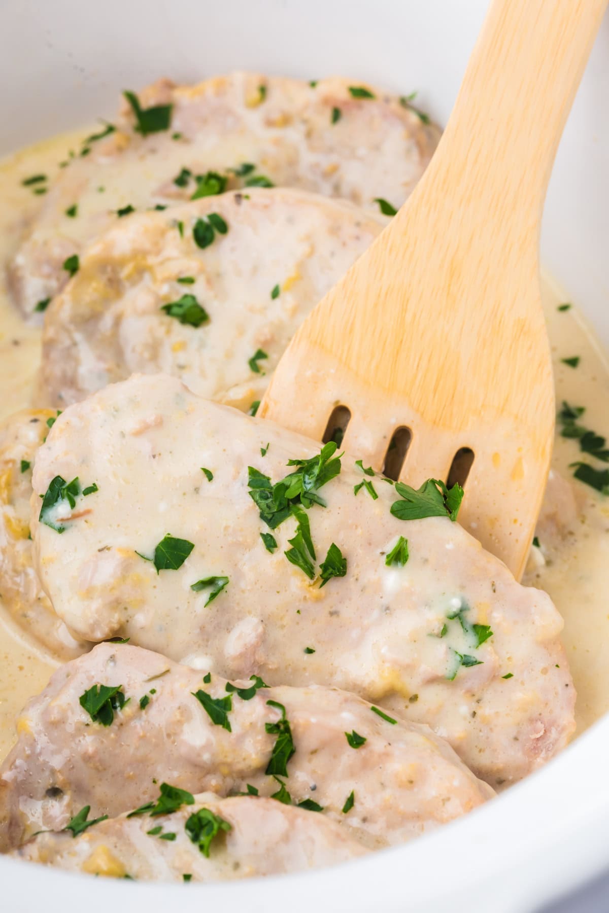 pork chops in a creamy ranch sauce being cooked in a crockpot with a wooden spoon taking one out.