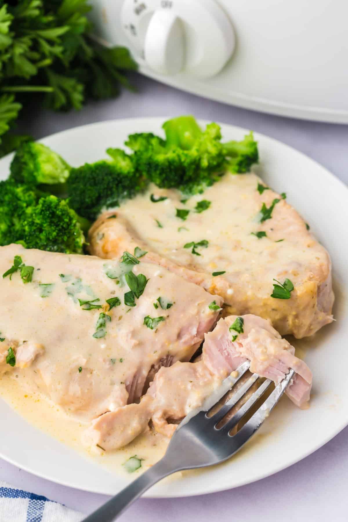 A fork taking a piece of Slow Cooker Creamy Ranch Pork Chops on a white plate with broccoli, in front of a crockpot.