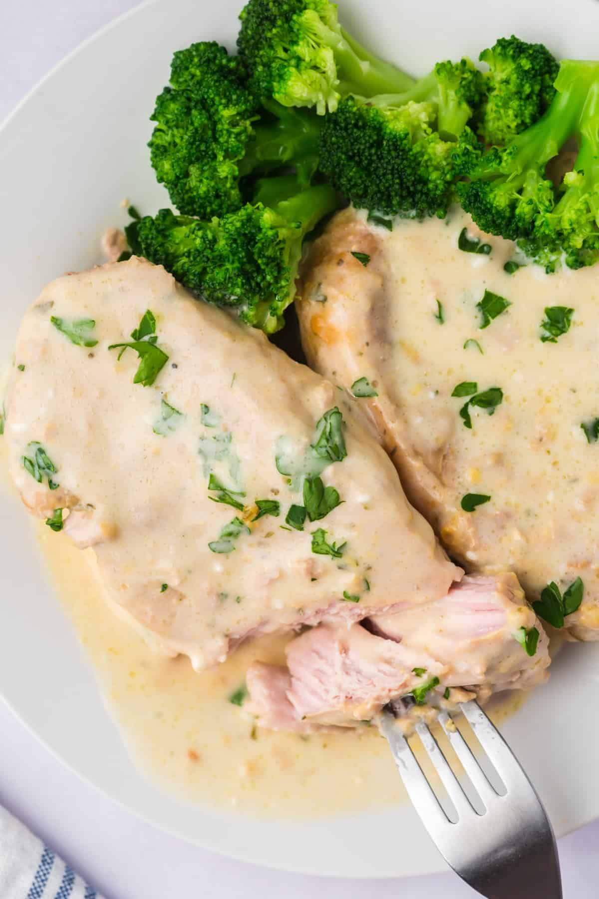 A fork taking a piece of Slow Cooker Creamy Ranch Pork Chops on a white plate with broccoli.