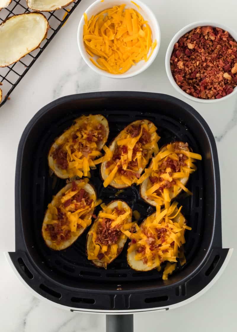 Potato Skins in an air fryer basket with bowls of shredded cheese and bacon beside it.