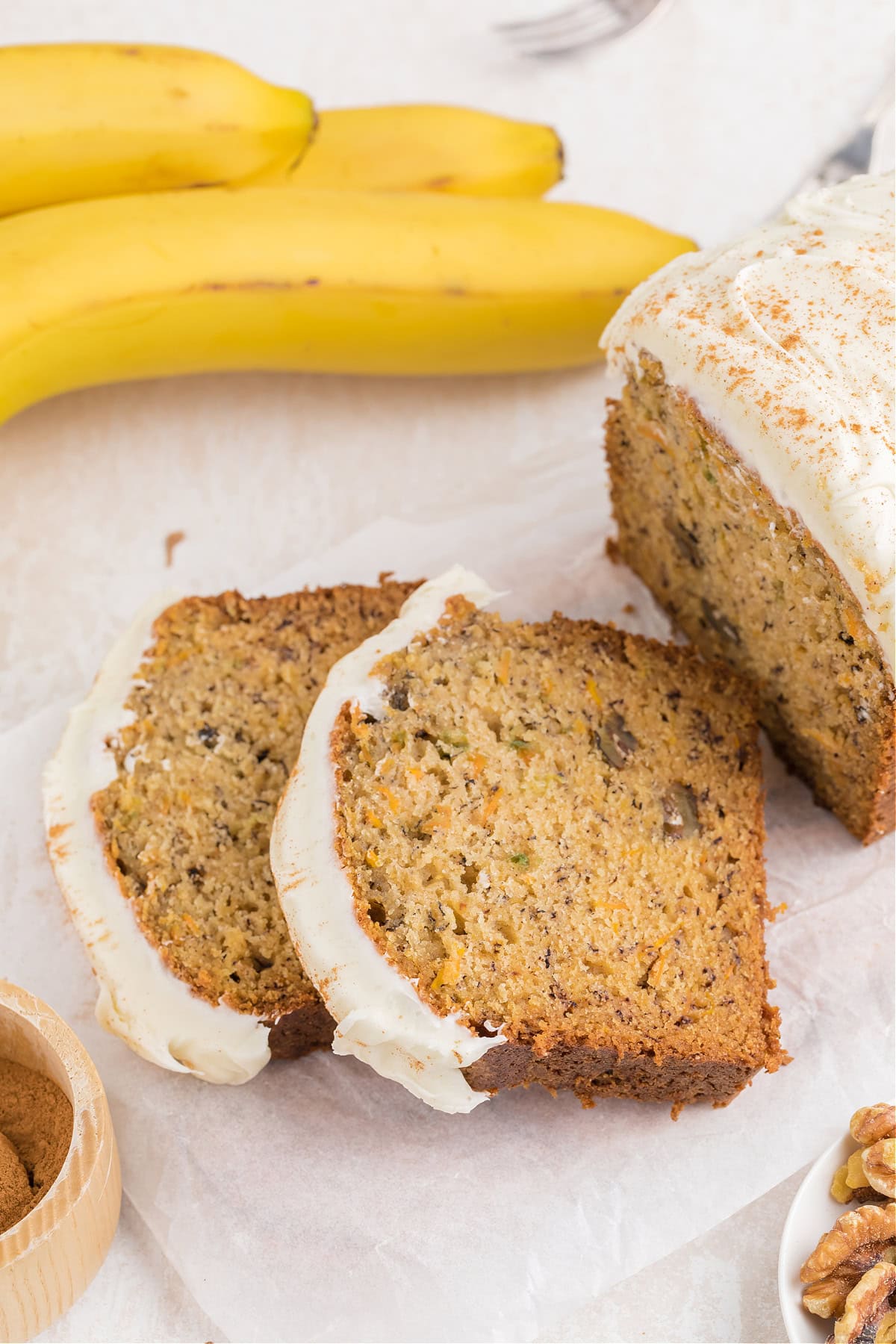 Sliced Carrot Cake Banana Bread on a table next to bananas and chopped walnuts.
