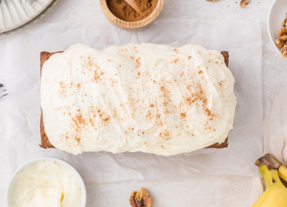 A whole frosted Carrot Banana Bread on a table next to bananas and walnuts.