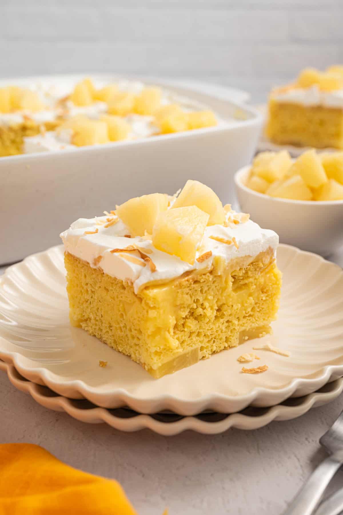 A slice of Pineapple Poke Cake on a round plate with pineapple chunks and toasted coconut on top.
