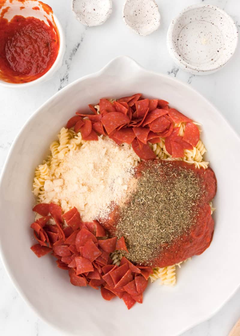 a bowl of ingredients to make pizza casserole including pasta, pepperoni, cheese and seasoning.
