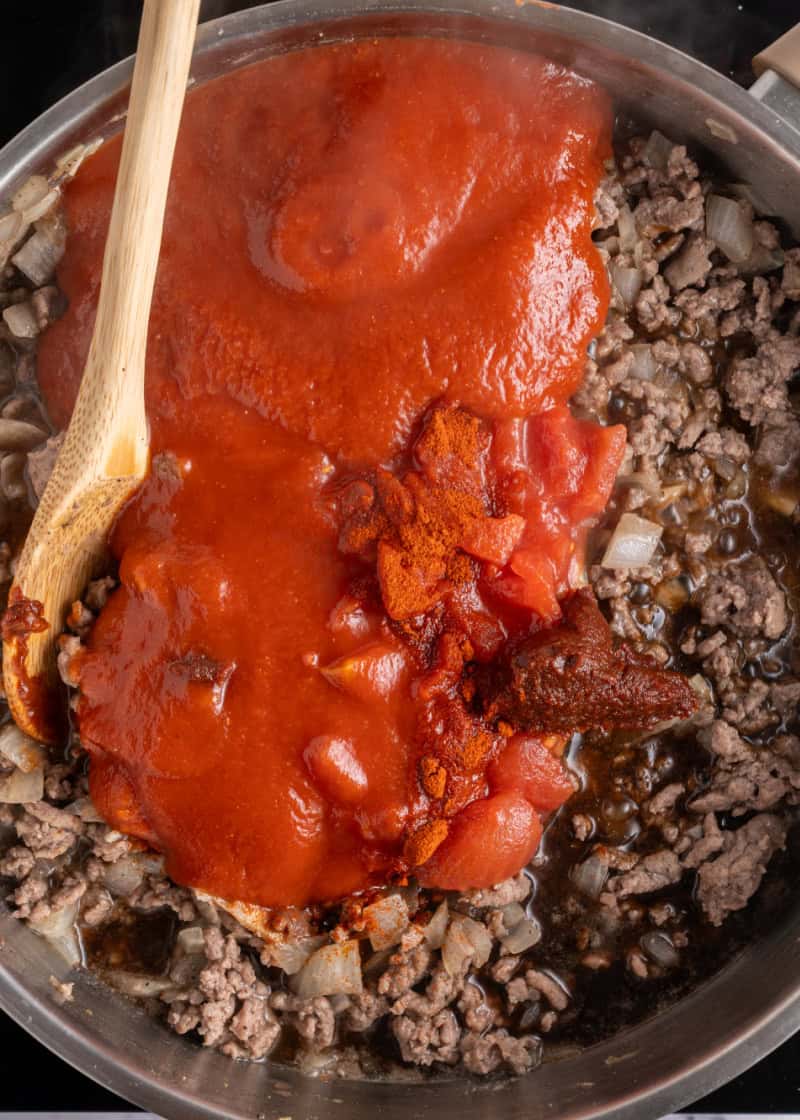 tomato sauces being added to cooked ground beef and onion in a frying pan.