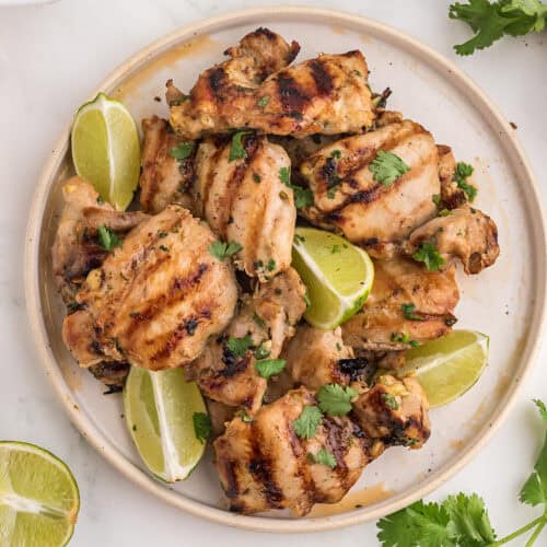 Pieces of Thai BBQ Chicken on a white plate with fresh cilantro and lime wedges.