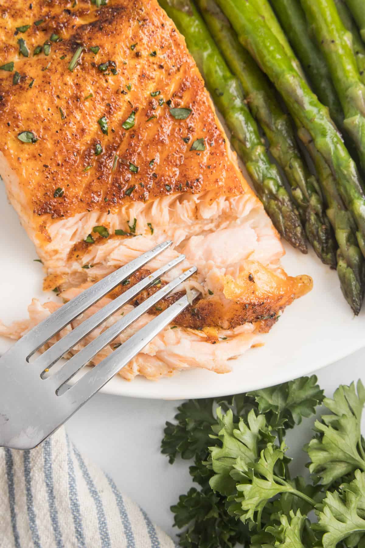 A fork taking a piece of salmon off a white plate with asparagus beside it.