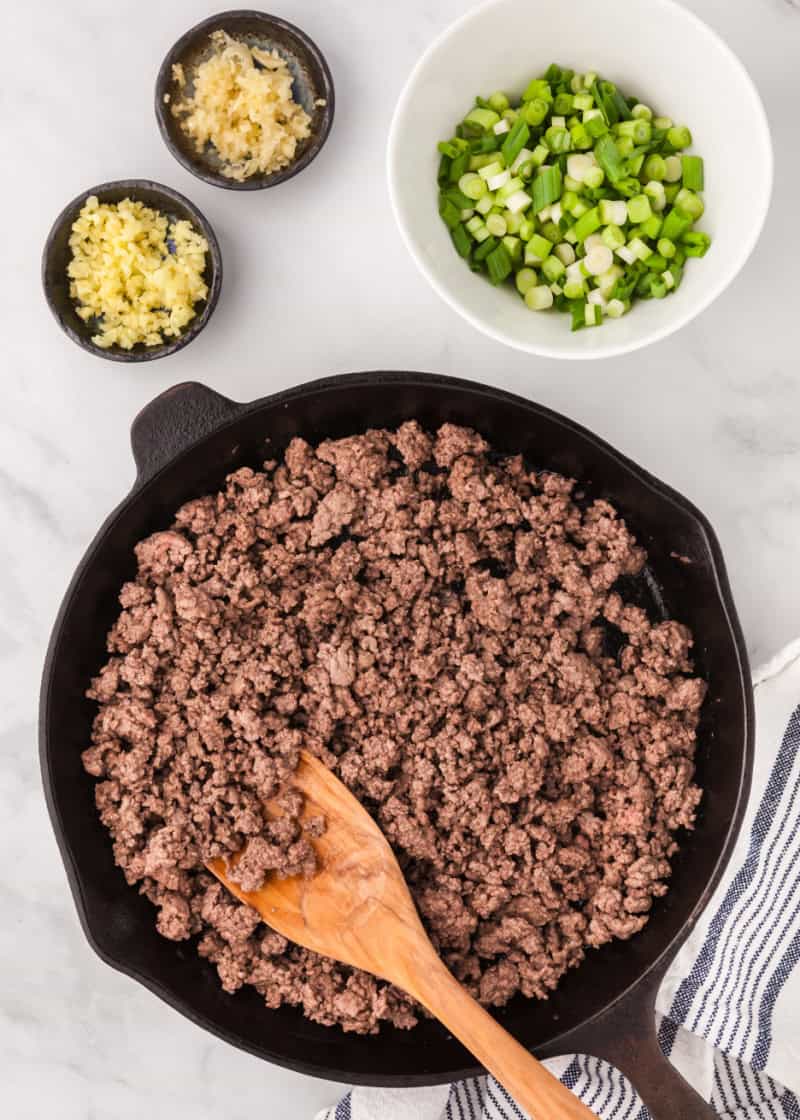 ground beef cooking in a skillet with bowls of green onion, garlic, and ginger beside it.