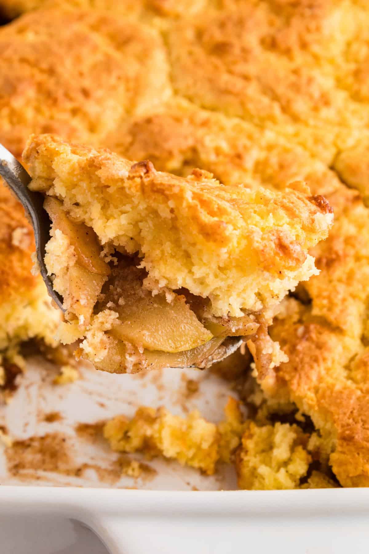 A spoon picking up cake mix apple cobbler out of a baking pan.